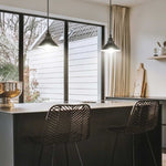 Modern kitchen interior with a dark island, two black wire bar stools, pendant lights, and a large window looking out onto a white house exterior.