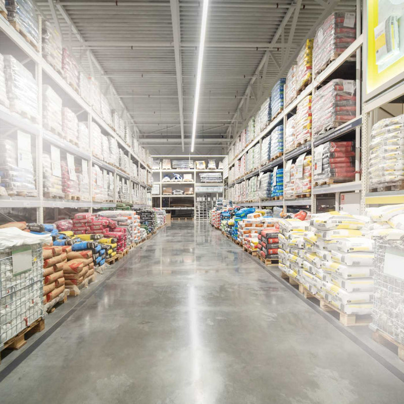 A long aisle in a large warehouse store, with tall shelving on both sides stocked with packaged goods and bags of materials. The floor is concrete and reflects overhead lighting.