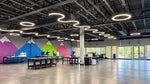 A wide interior view of a modern office or studio space with a high ceiling, black metal trusses, wavy ring LED light fixtures, long work tables, and colorful mountain wall murals in purple, blue, green, and pink. Large windows on the right let in natural light.