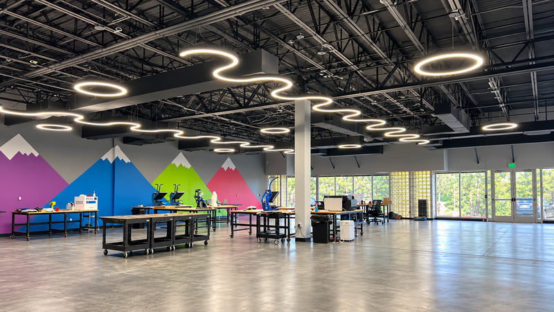 A wide interior view of a modern office or studio space with a high ceiling, black metal trusses, wavy ring LED light fixtures, long work tables, and colorful mountain wall murals in purple, blue, green, and pink. Large windows on the right let in natural light.