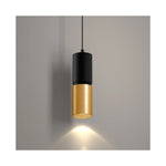 A modern cylindrical pendant light with a matte black upper section and a brushed gold lower shade, suspended by a thin black cord against a neutral gray wall with a warm light beam casting downward.