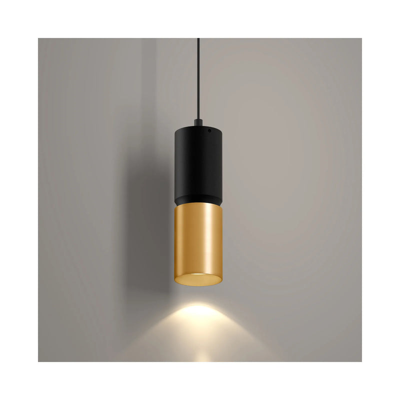 A modern cylindrical pendant light with a matte black upper section and a brushed gold lower shade, suspended by a thin black cord against a neutral gray wall with a warm light beam casting downward.