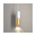 A modern pendant light with a white upper cylinder and a brushed gold lower cylinder, shown illuminated against a neutral gray wall.