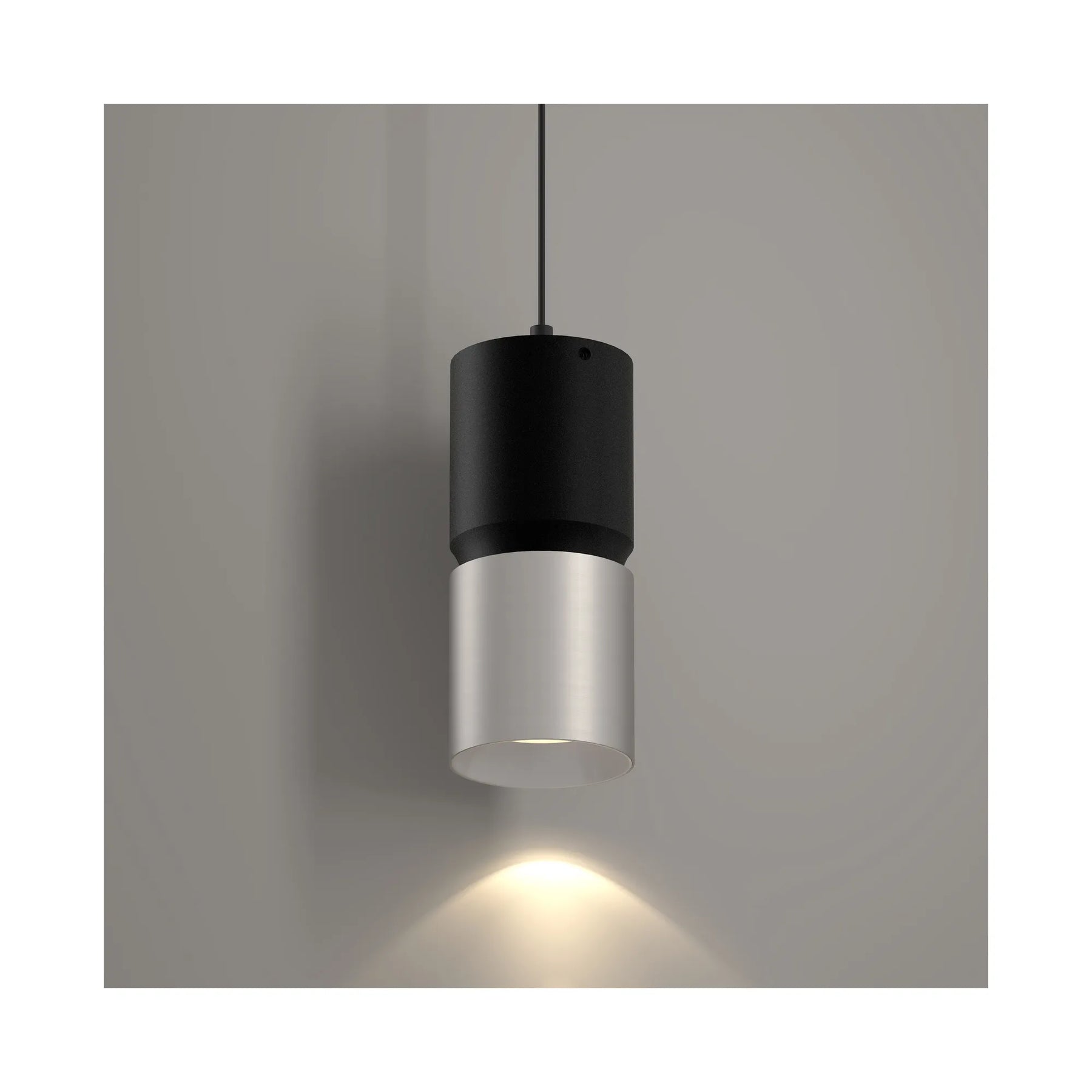 A modern cylindrical pendant light with a matte black upper section and a brushed nickel lower shade, suspended against a neutral gray wall with a warm light beam casting downward.