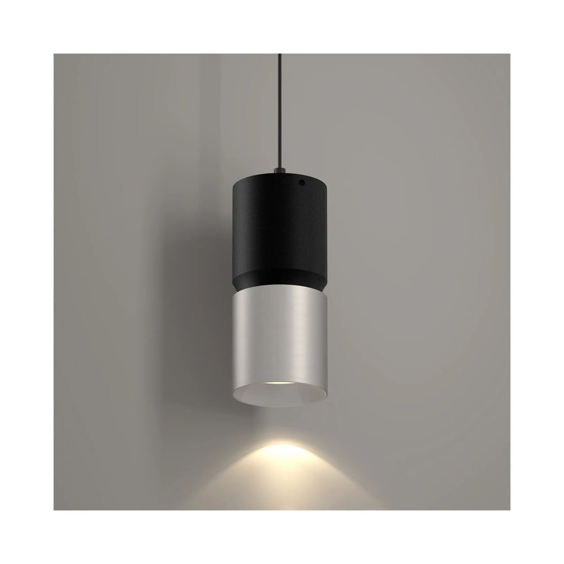 A modern cylindrical pendant light with a matte black upper section and a brushed nickel lower shade, suspended against a neutral gray wall with a warm light beam casting downward.