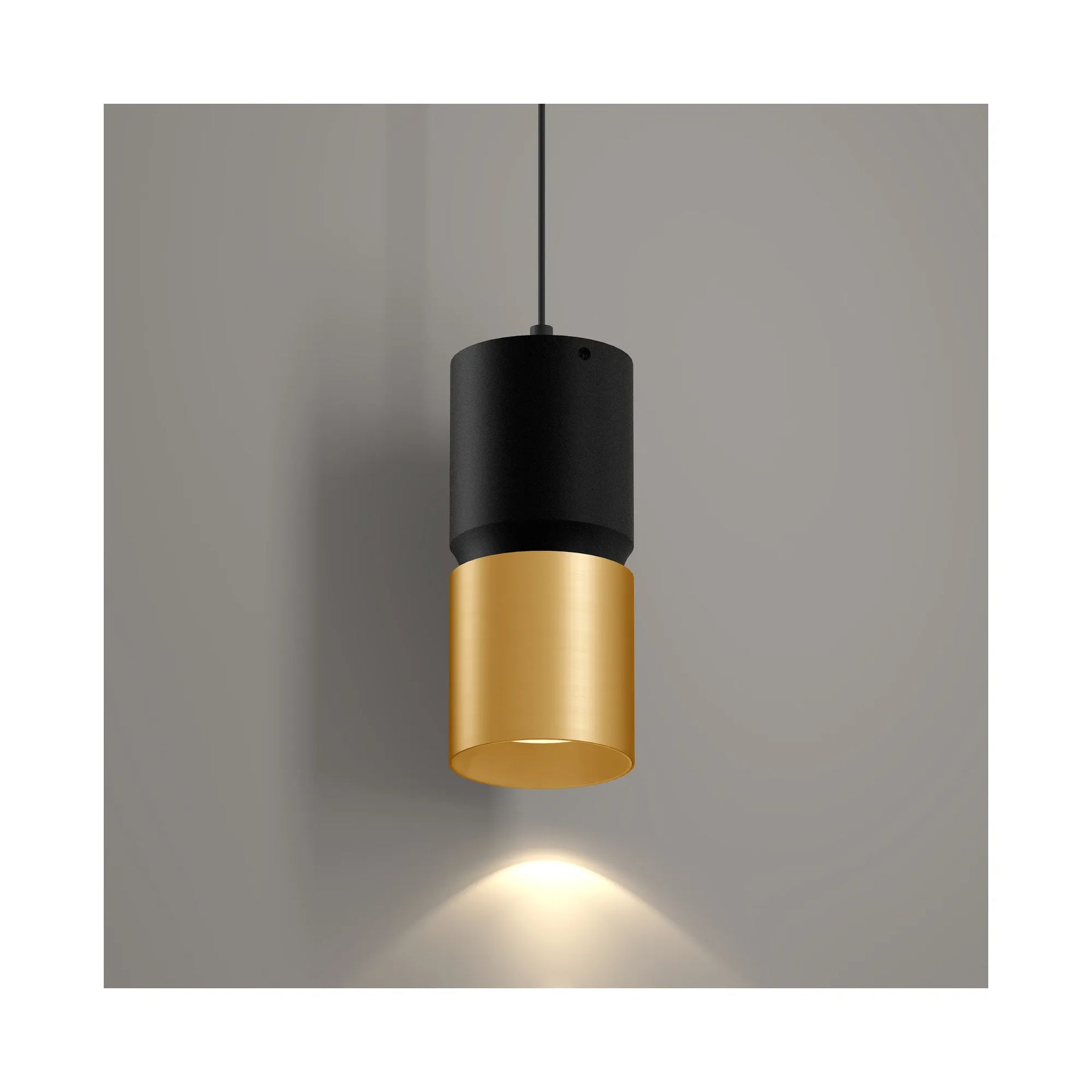 A modern cylindrical pendant light with a matte black upper section and a brushed gold lower shade, suspended by a thin black cord against a neutral gray wall with a warm light beam casting downward.