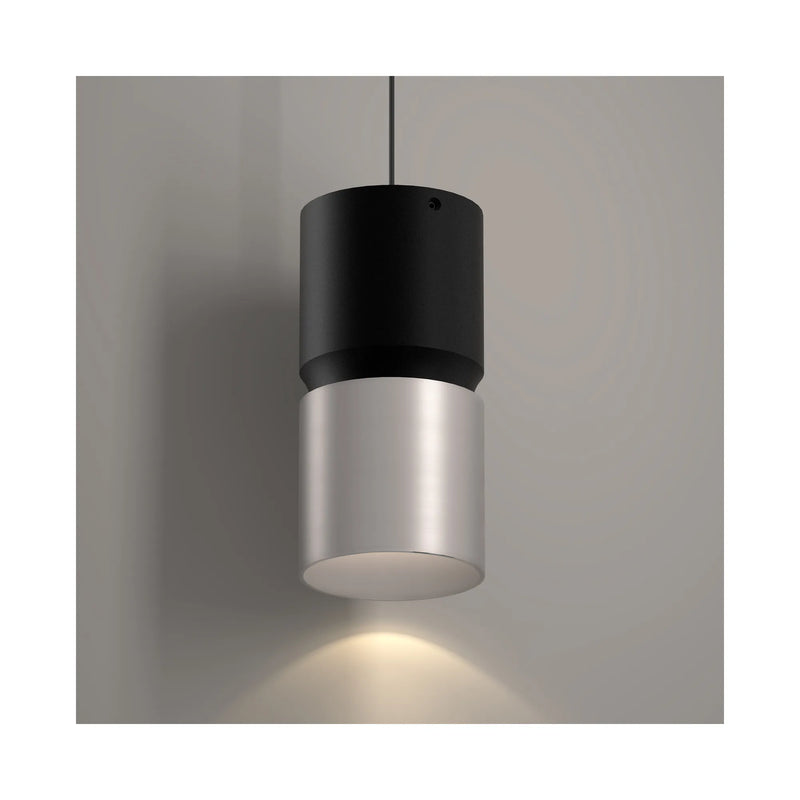 A modern cylindrical pendant light with a matte black top section and a brushed nickel metal lower shade, suspended against a neutral gray wall with a warm light glowing downward.