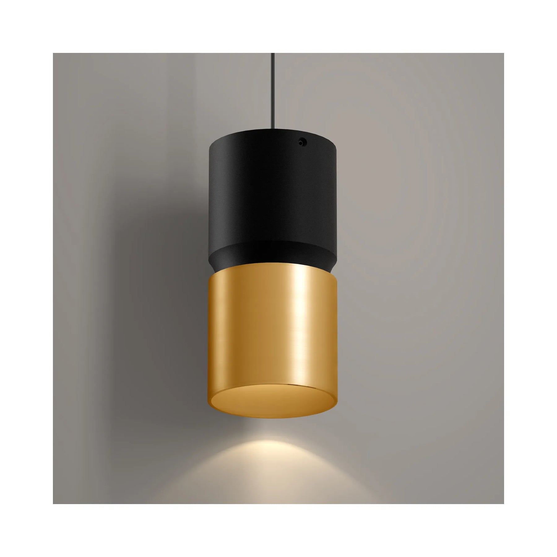 A modern cylindrical pendant light with a matte black upper shade and a brushed gold lower shade, suspended against a neutral gray wall with a warm light glowing downward.