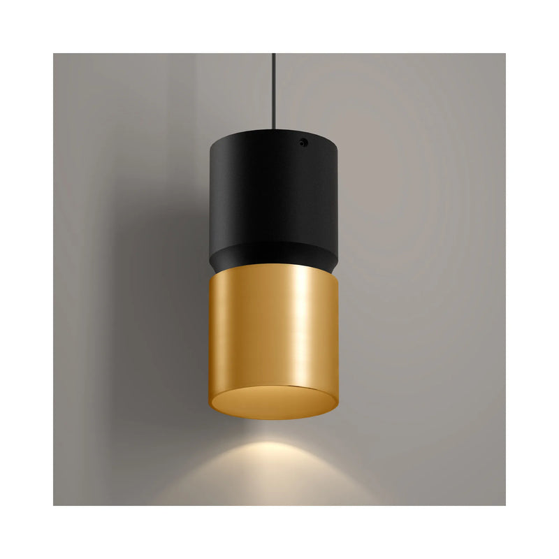 A modern cylindrical pendant light with a matte black upper shade and a brushed gold lower shade, suspended against a neutral gray wall with a warm light glowing downward.