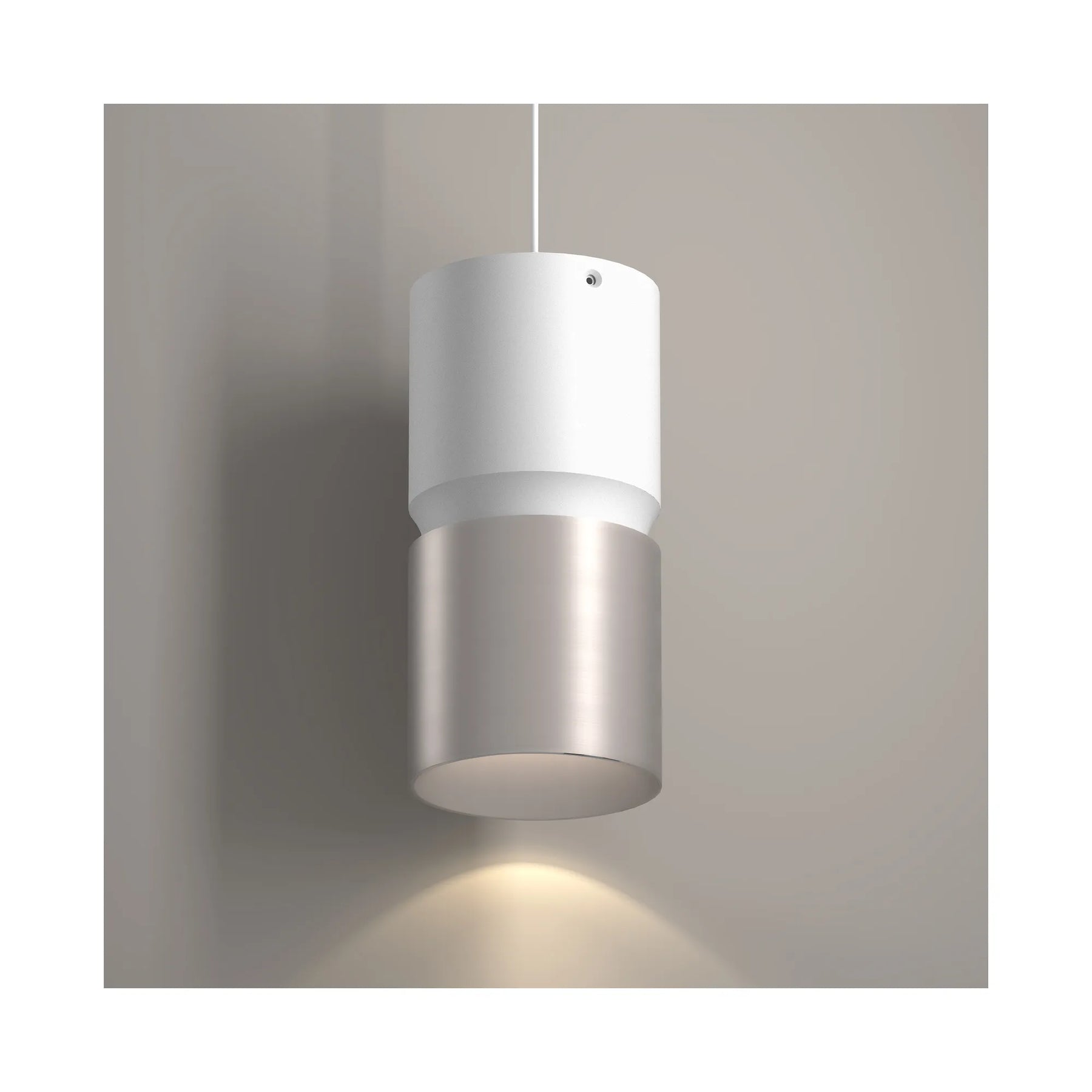 A modern cylindrical pendant light with a matte white upper section and a brushed nickel lower section, shown hanging against a neutral gray wall with a warm light emitted below.