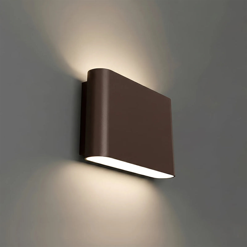 A modern rectangular wall sconce light fixture in a bronze finish, mounted on a gray wall and casting a warm light downward.