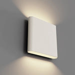 A modern square wall sconce light fixture in a warm off-white finish, shown mounted on a gray wall with warm light projected upwards and downwards.
