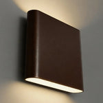 A modern square wall sconce light fixture in a bronze finish, mounted on a gray wall and casting a warm light downward.