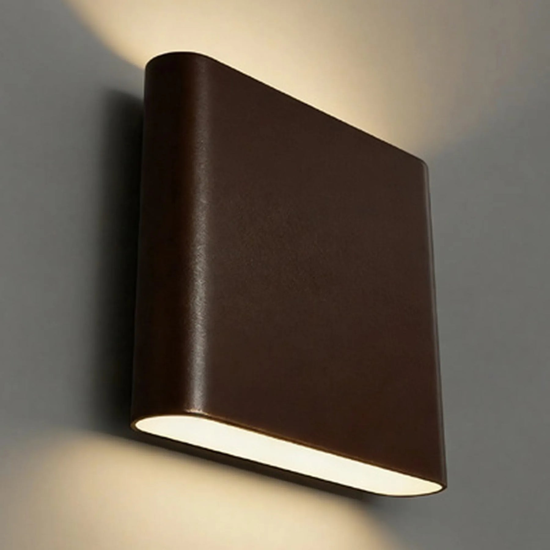 A modern square wall sconce light fixture in a bronze finish, mounted on a gray wall and casting a warm light downward.