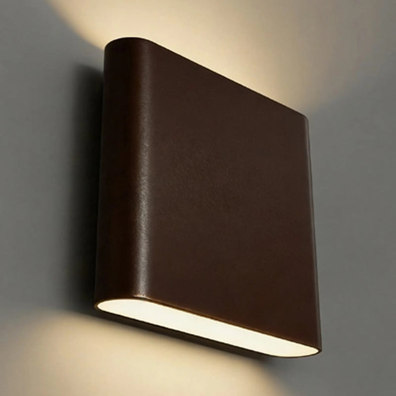 A modern square wall sconce light fixture in a bronze finish, mounted on a gray wall and casting a warm light downward.